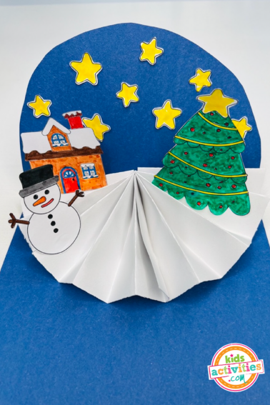 Image shows a finished Christmas house craft: it's a construction paper craft with a blue background, stars, a Christmas tree, a snowman and a house. From Kids Activities Blog