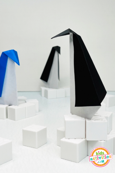 Image shows 3 origami penguin crafts standing on their own on white paper blocks. Tutorial from Kids Activities Blog